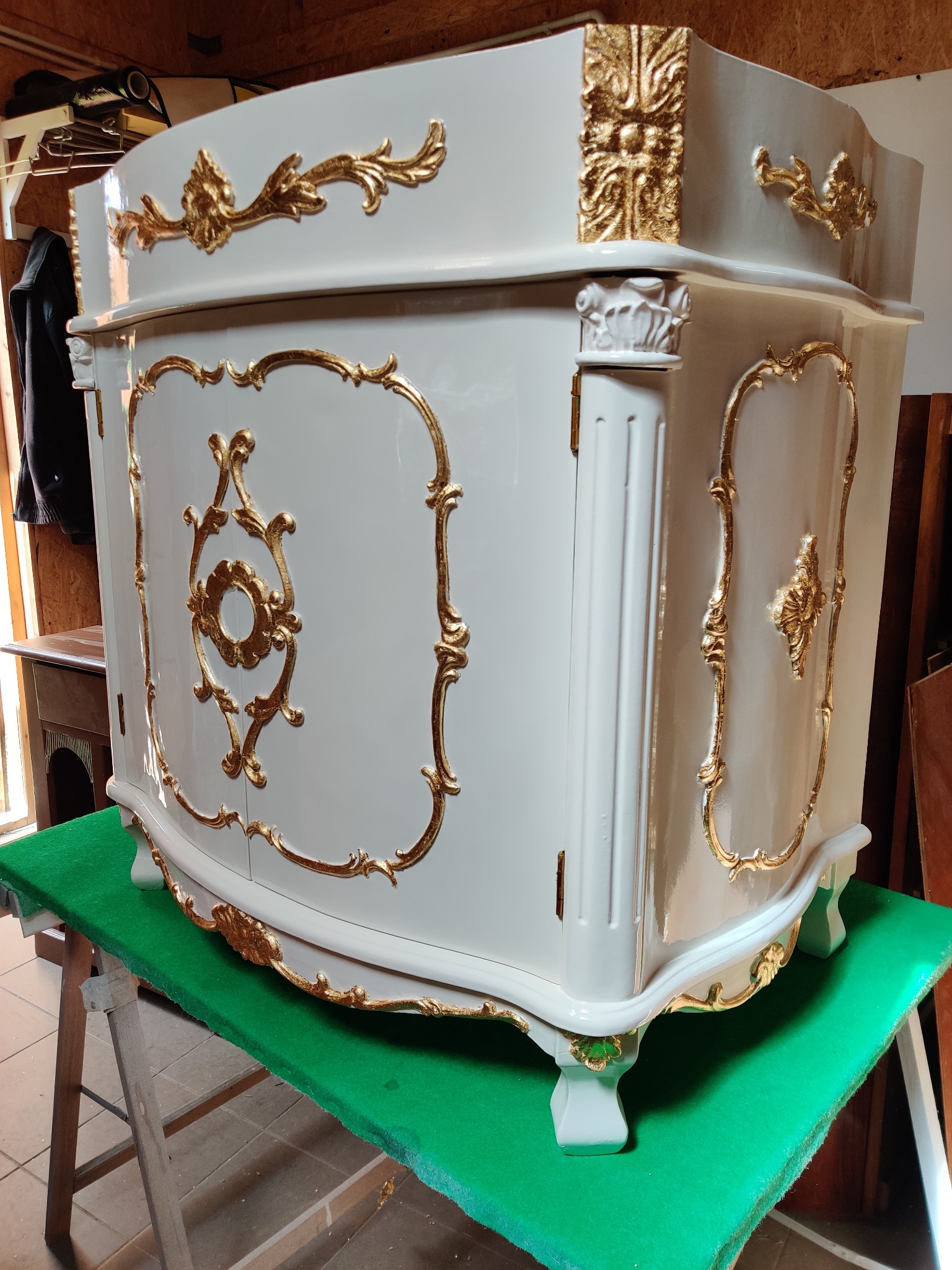 Biała, lakierowana komoda w stylu Ludwika z bogatymi, złoconymi zdobieniami, ustawiona na zielonym materiale na drewnianym koźle.