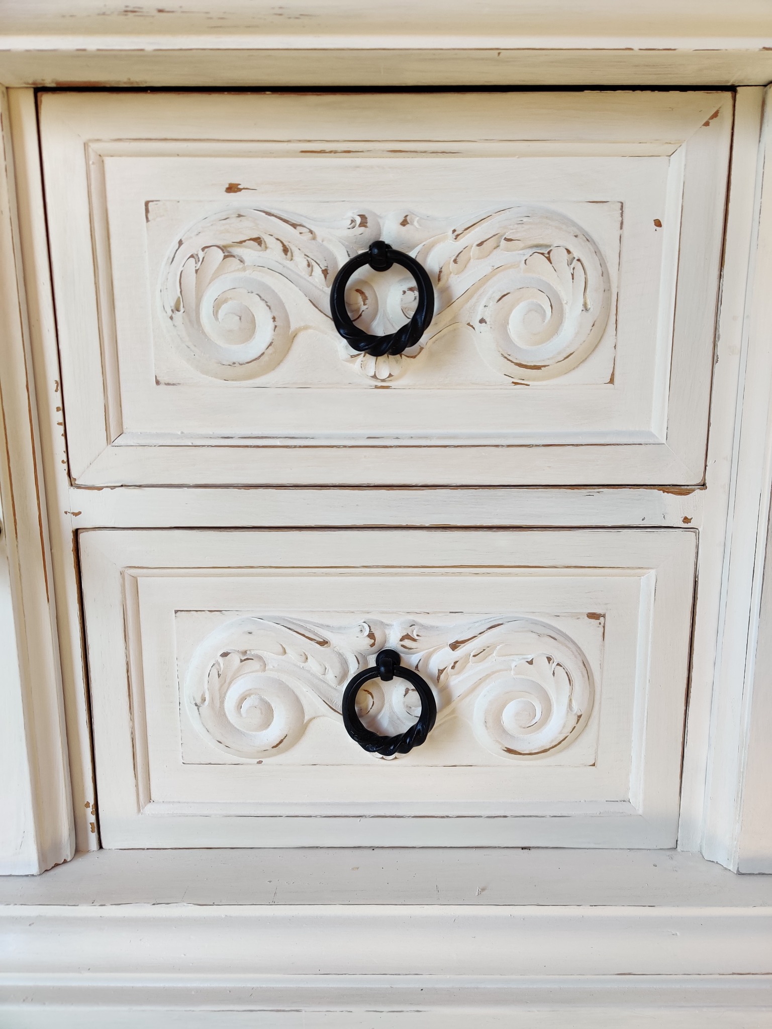 Dwa białe fronty szuflad z dekoracyjnymi, wyżłobionymi ornamentami i czarnymi, okrągłymi uchwytami, stylizowane na postarzane.