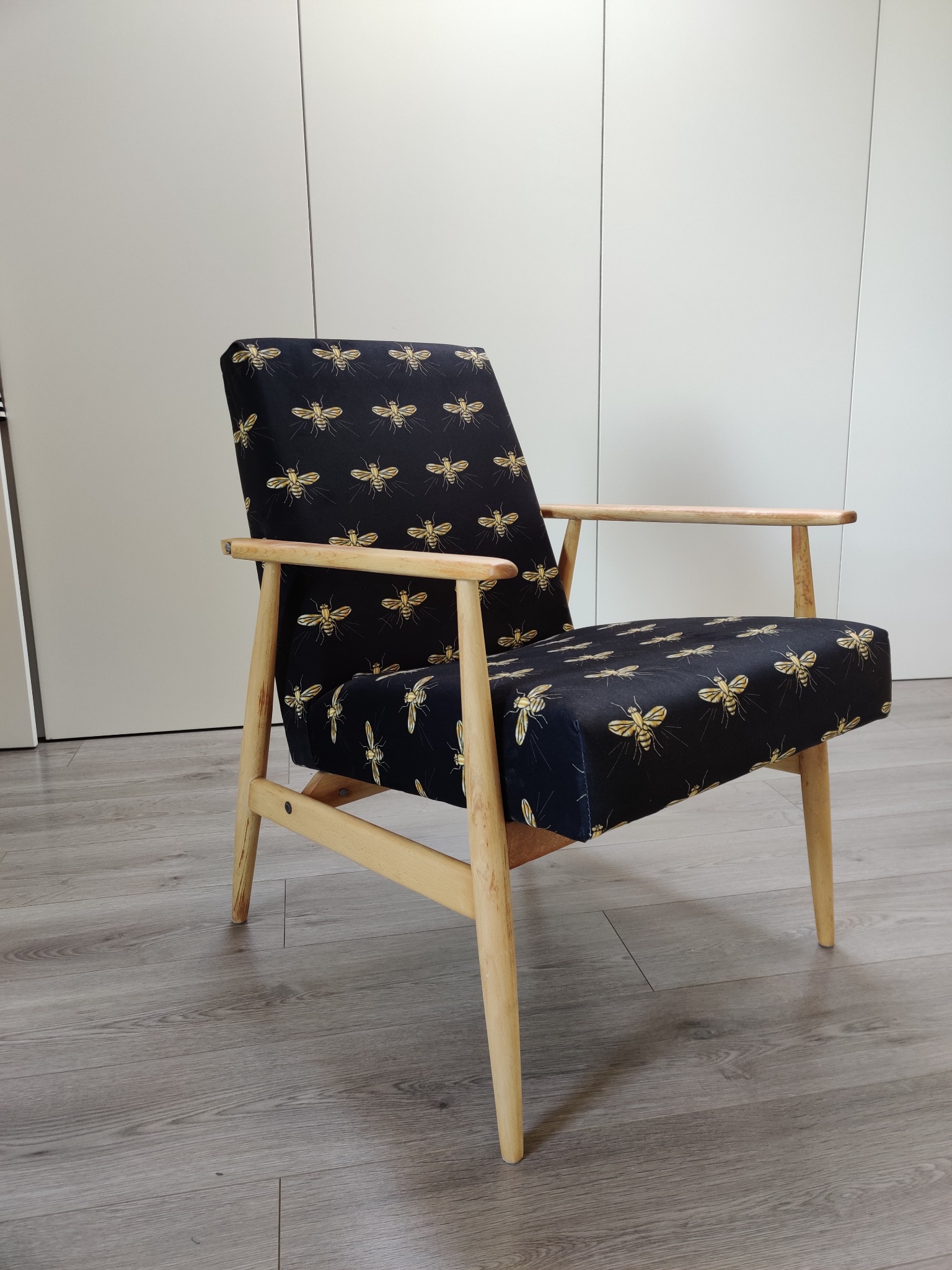 Fotel w stylu retro z jasnego drewna, obity czarną tkaniną w złote pszczoły, na tle białych szaf i szarej podłogi.