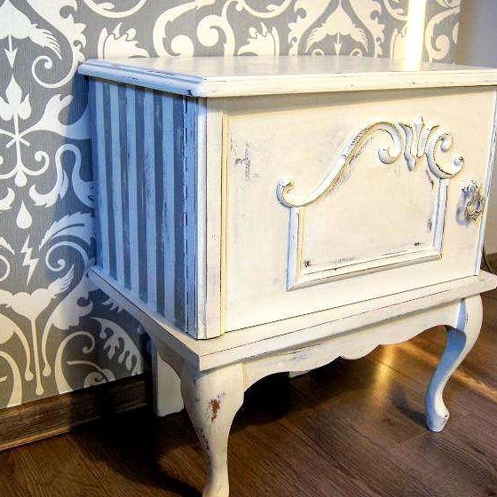 Biała, odnowiona szafka nocna w stylu shabby chic z szarymi paskami na boku, z dekoracyjnym ornamentem na drzwiczkach, stojąca na drewnianej podłodze na tle wzorzystej tapety.