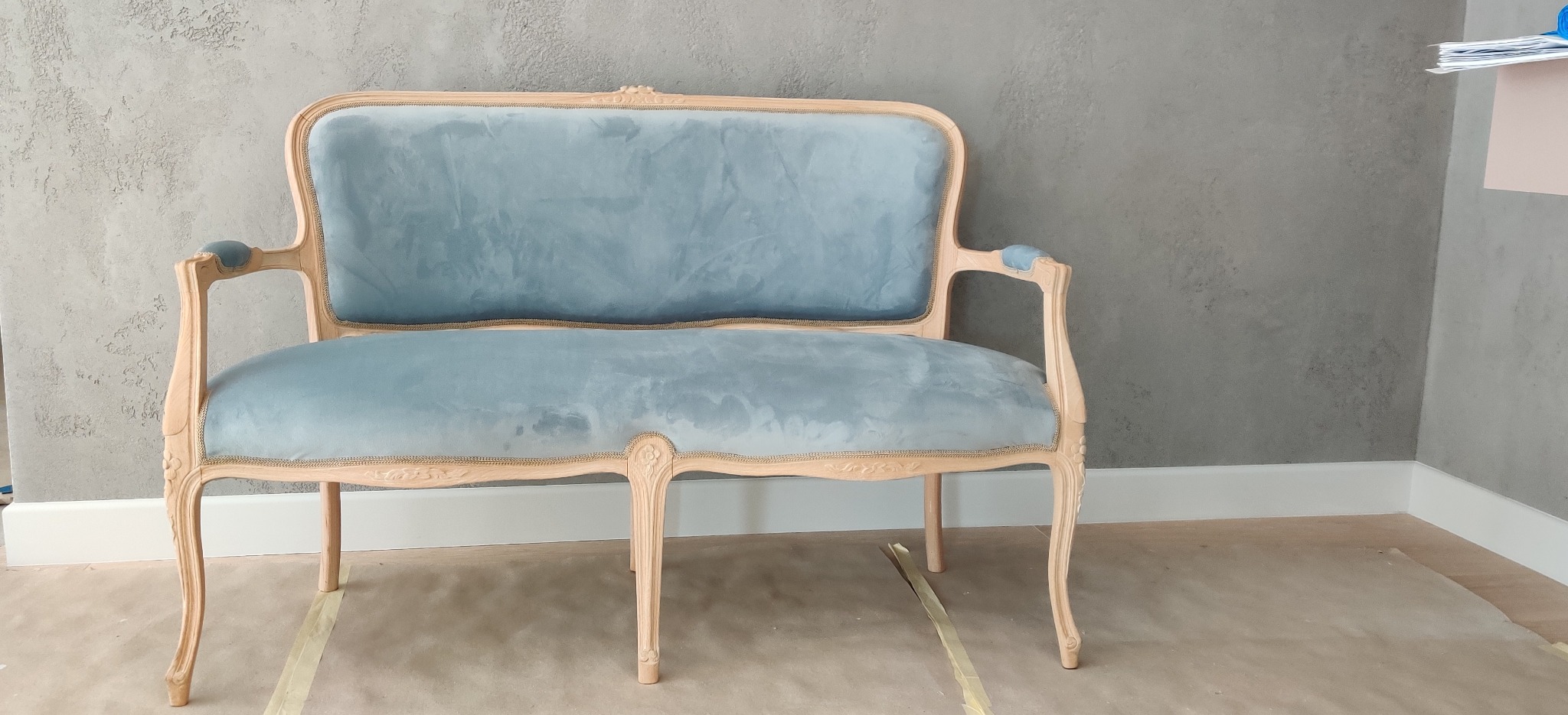 Dwuosobowa kanapa w stylu Ludwika XVI z jasnym drewnianym szkieletem i nową, błękitną welurową tapicerką, stojąca na podłodze zabezpieczonej taśmą malarską.