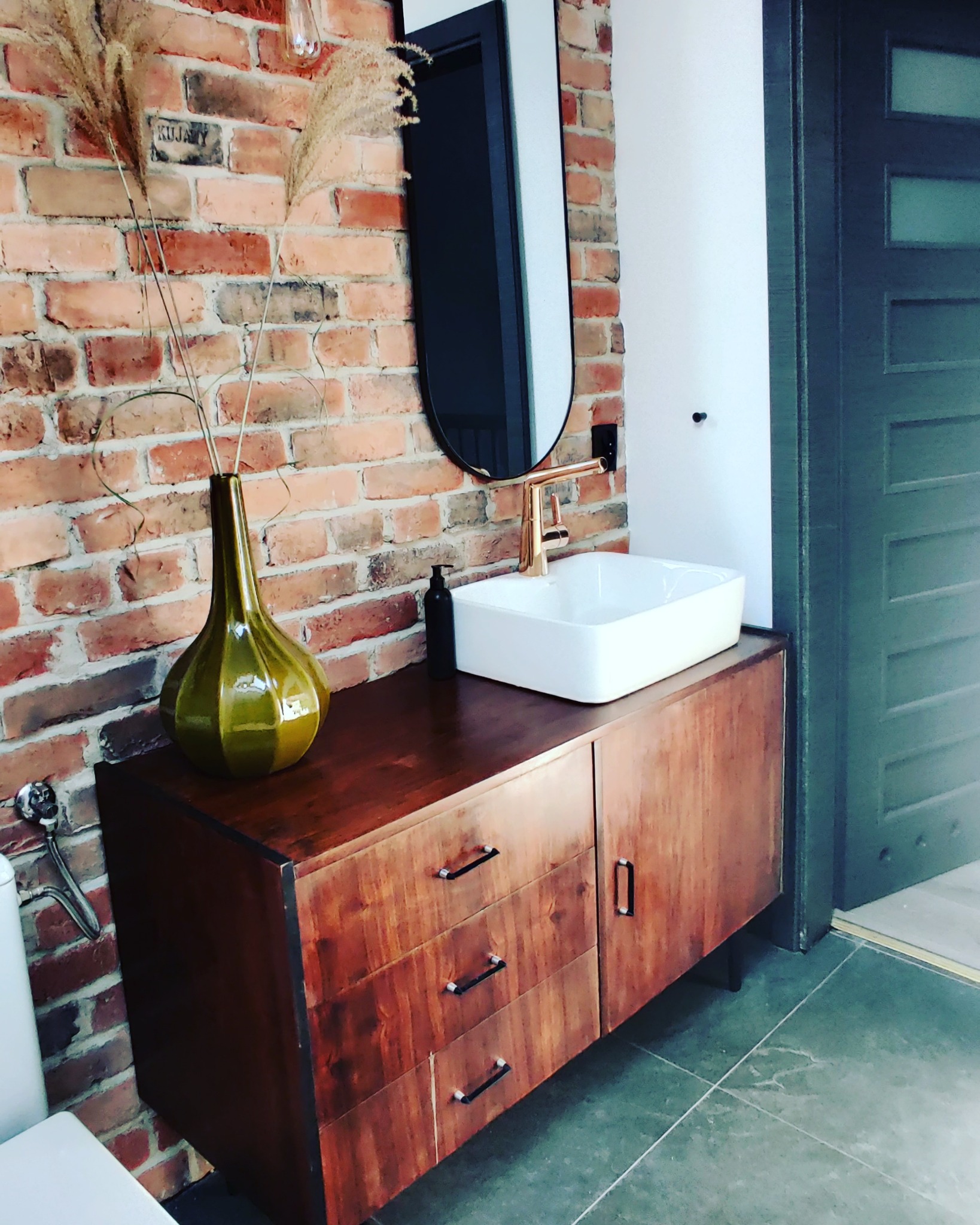 Nowoczesna łazienka z umywalką nablatową, baterią w kolorze miedzi, lustrem w czarnej ramie i drewnianą szafką, na tle ściany z czerwonej cegły i dekoracyjnym wazonem z suszonymi trawami.