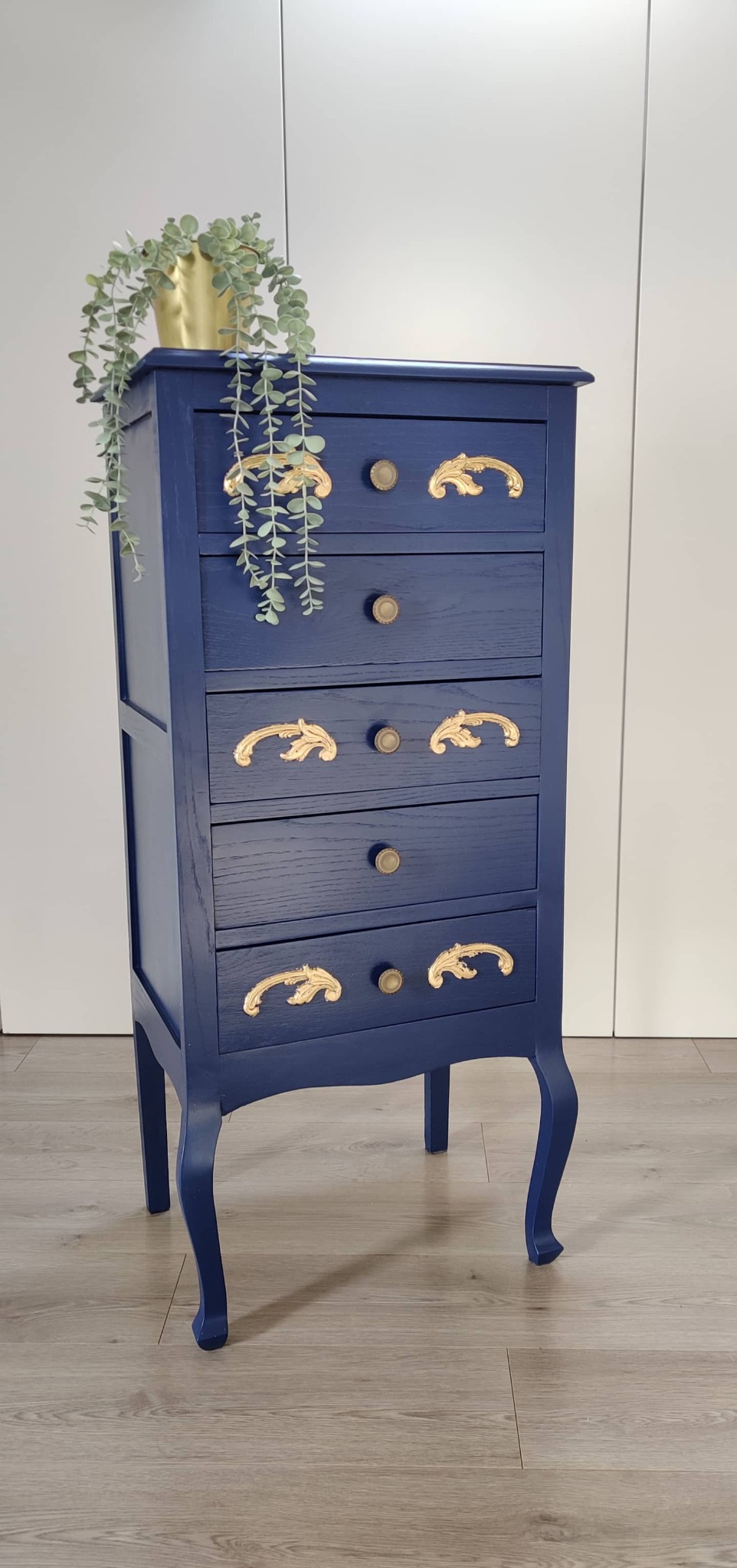 Odnowiona, granatowa komoda z pięcioma szufladami, zdobiona złotymi ornamentami i okrągłymi gałkami, na blacie doniczka ze zwisającą rośliną, na tle białej ściany i jasnej podłogi.