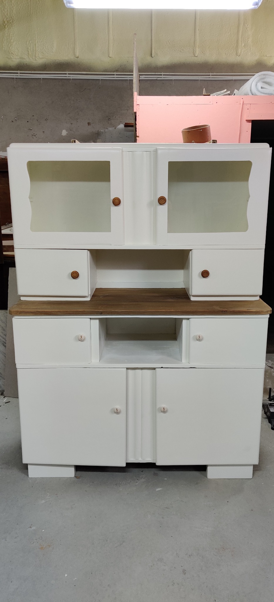 Odrestaurowany, dwuczęściowy kredens w stylu art deco, z białymi frontami, drewnianym blatem i okrągłymi uchwytami, stojący na betonowej podłodze w warsztacie.