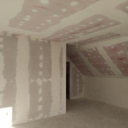 Usługi remontowo budowlane - Pokój na poddaszu w trakcie przygotowania ścian i sufitu do malowania, widoczne płyty gipsowo-kartonowe z miejscami szpachlowania, surowy beton na podłodze, wiszący kabel.