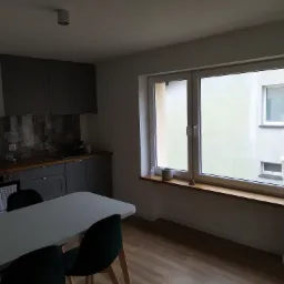 Nowoczesna kuchnia z szarymi szafkami, wzorzystą ścianą nad blatem i dużym oknem z drewnianym parapetem, na którym stoją dekoracje.