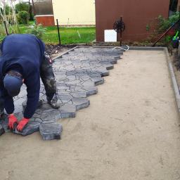 Usługi remontowo budowlane - Układanie nieregularnej kostki brukowej na przygotowanym podłożu w Kołobrzegu, widoczne czerwone rękawice robocze i częściowo ułożona powierzchnia.