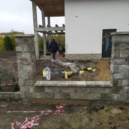 Usługi remontowo budowlane - Budowa ogrodzenia z szarych, betonowych bloczków przy nowo wybudowanym domu z białymi ścianami i drewnianą konstrukcją tarasu; widoczne narzędzia i materiały budowlane.