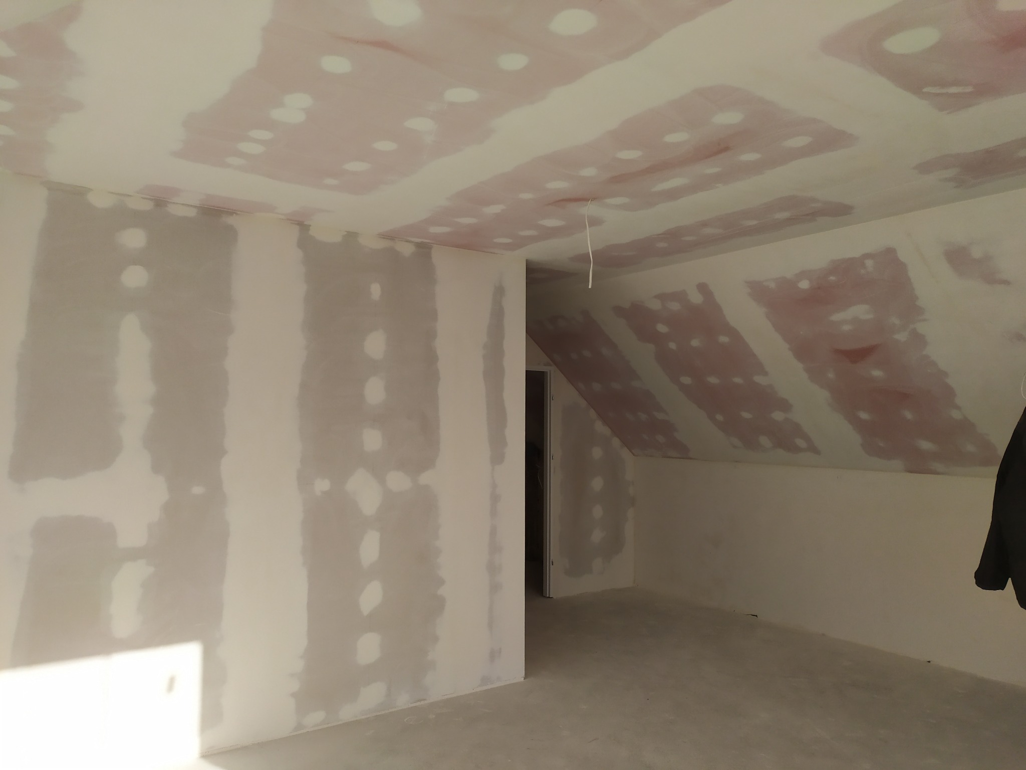 Pokój na poddaszu w trakcie przygotowania ścian i sufitu do malowania, widoczne płyty gipsowo-kartonowe z miejscami szpachlowania, surowy beton na podłodze, wiszący kabel.