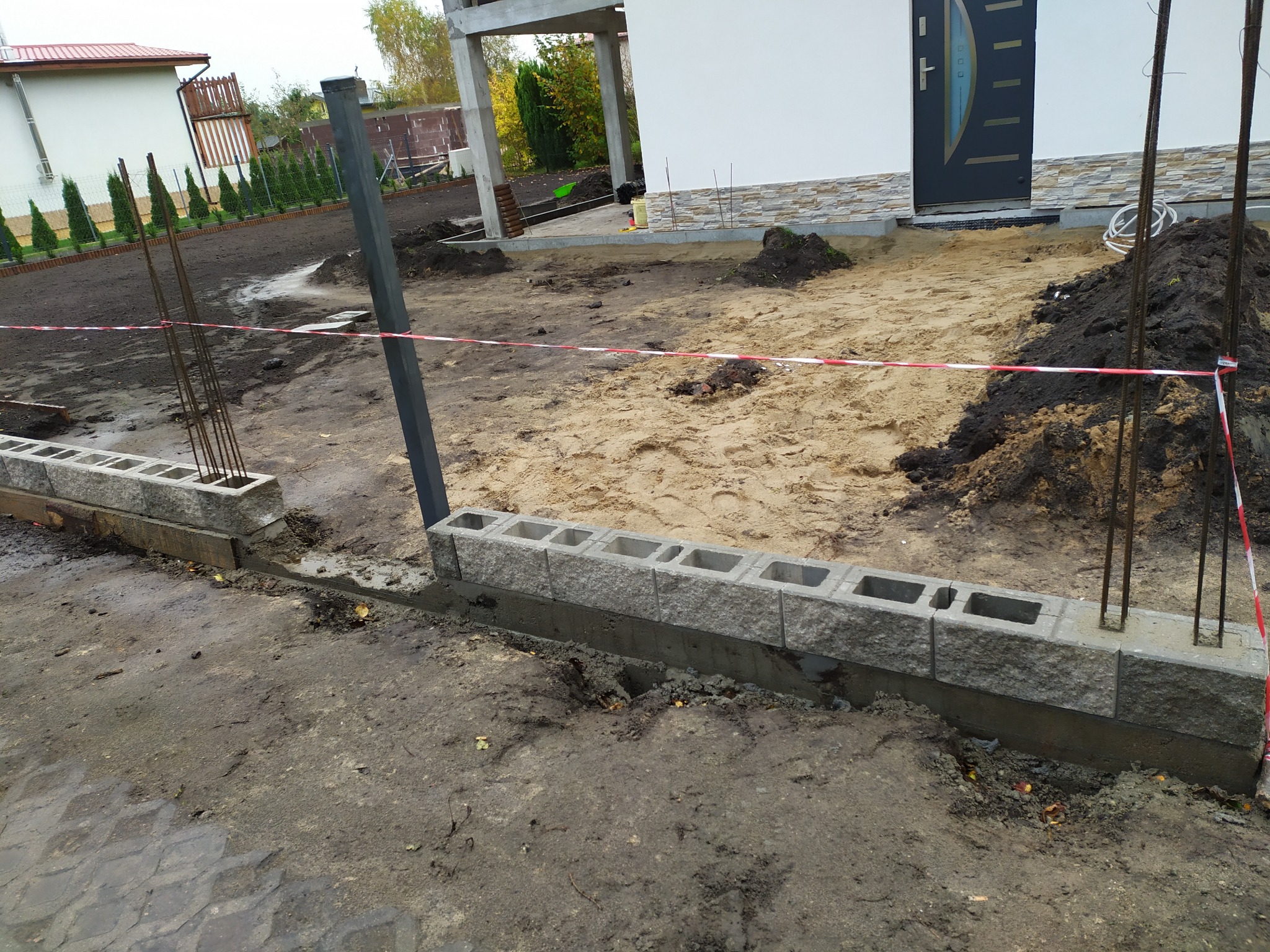 Wznoszenie fundamentu z betonowych bloczków na posesji, widoczne pręty zbrojeniowe i taśma ostrzegawcza.