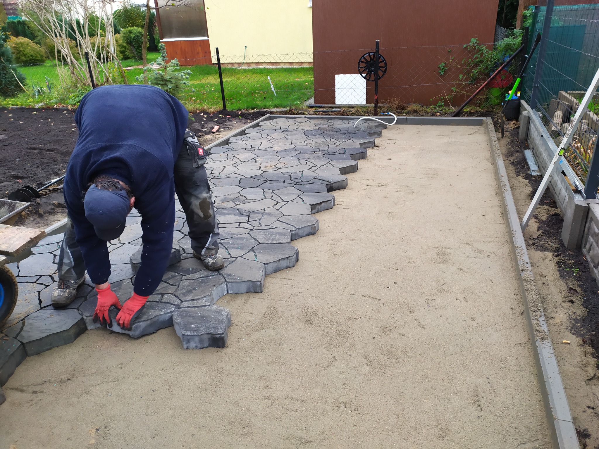 Układanie nieregularnej kostki brukowej na przygotowanym podłożu w Kołobrzegu, widoczne czerwone rękawice robocze i częściowo ułożona powierzchnia.