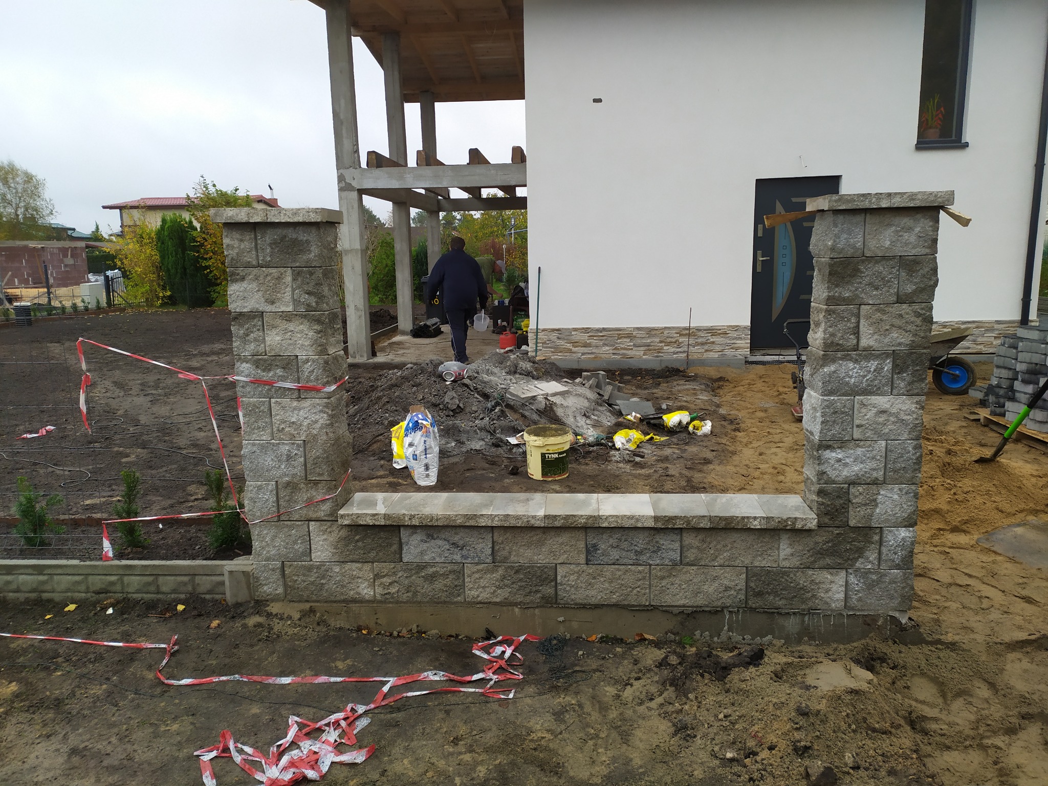 Budowa ogrodzenia z szarych, betonowych bloczków przy nowo wybudowanym domu z białymi ścianami i drewnianą konstrukcją tarasu; widoczne narzędzia i materiały budowlane.