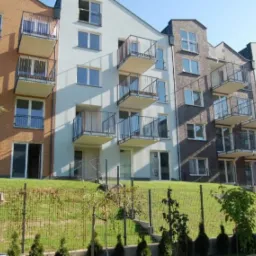 Nowoczesny budynek wielorodzinny z balkonami, elewacja z cegły i białego tynku, widok z poziomu trawnika, z niskim ogrodzeniem i młodymi drzewkami na pierwszym planie.