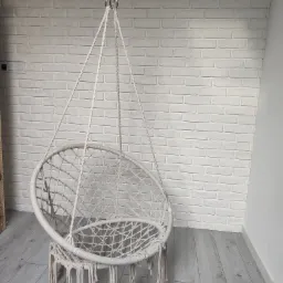 Beżowe, wiszące krzesło hamakowe z frędzlami, zawieszone na metalowym łańcuchu, na tle ściany z białej cegły i szarej podłogi z paneli.