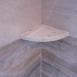 Narożna, marmurowa półka w kabinie prysznicowej, z szarymi płytkami imitującymi drewno ułożonymi w jodełkę na dole ściany i gładkimi, szarymi płytkami u góry.