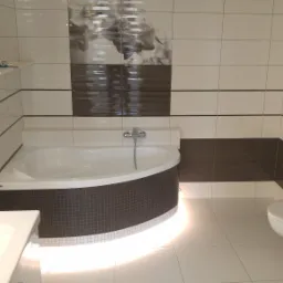 Nowoczesna łazienka z narożną wanną obudowaną mozaiką, białą ceramiką i brązowo-białymi płytkami, dekorowana motywem kwiatowym na ścianie.