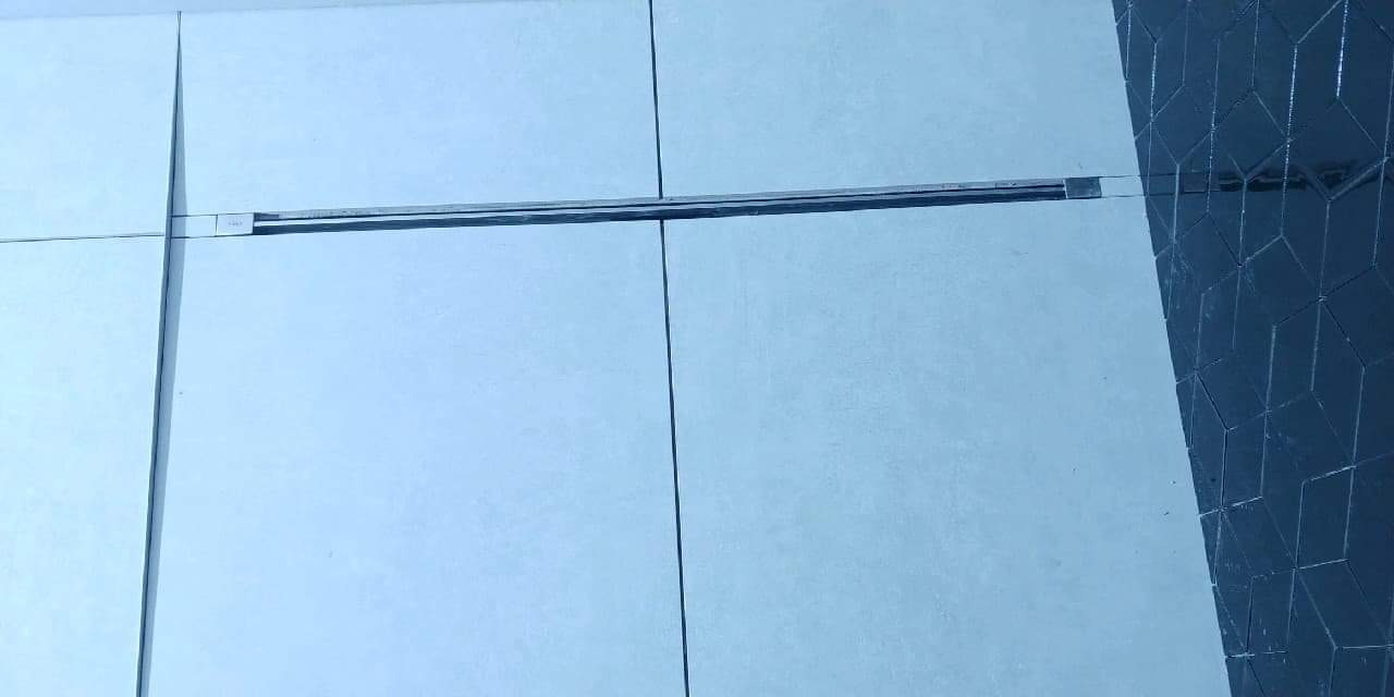 Jasne, niemal białe, kwadratowe płytki z wąskimi fugami i metalową listwą dekoracyjną, sąsiadujące z ciemnymi, trójwymiarowymi płytkami o wzorze kostki.