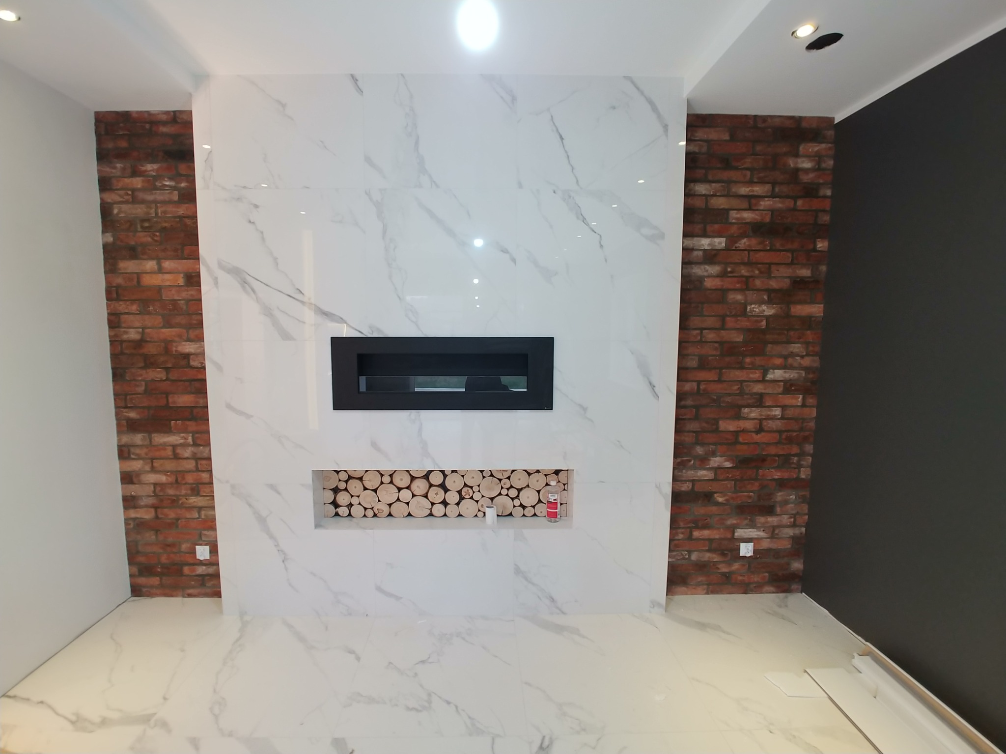 Wykończony salon z kominkiem obudowanym białymi płytami imitującymi marmur, z wnęką wypełnioną drewnem i ścianami akcentowanymi czerwoną cegłą.