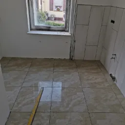 Wnętrze łazienki w trakcie prac wykończeniowych, beżowe płytki podłogowe ułożone z krzyżykami dystansowymi, częściowo położone białe płytki ścienne, poziomica leżąca na podłodze, widok przez okno...