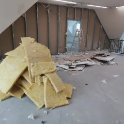 Poddasze w trakcie ocieplania: widoczna stelaż metalowy, płyty karton gips do montażu, stos wełny mineralnej i rozrzucone fragmenty materiałów izolacyjnych, drabina w tle.