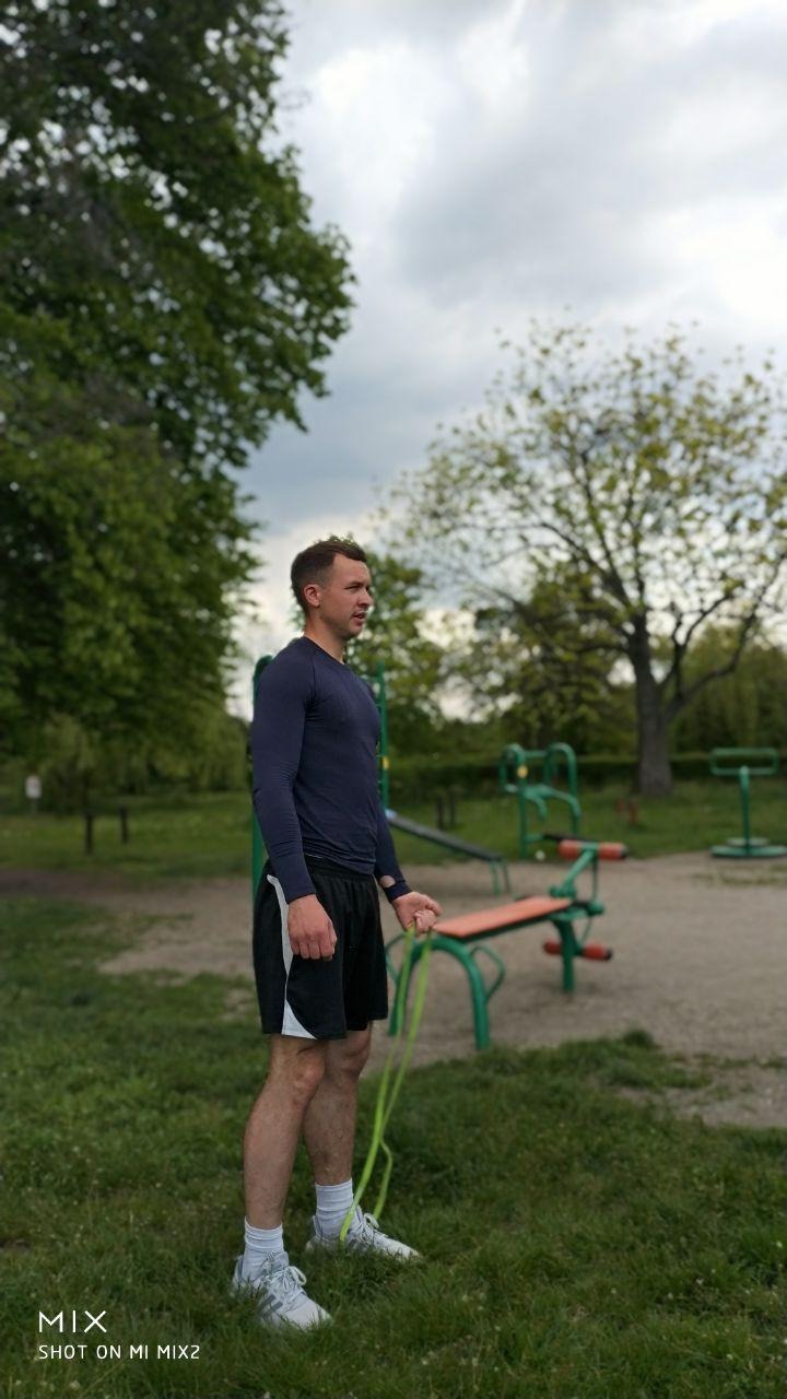 Mężczyzna w sportowym stroju, trzymający zieloną gumę do ćwiczeń na tle plenerowej siłowni w parku, pochmurne niebo.