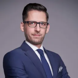 Portret mężczyzny w okularach i niebieskim garniturze, biała koszula i granatowy krawat, ciemne włosy zaczesane do tyłu, szara ściana w tle, kieszonka z ozdobną poszetką.