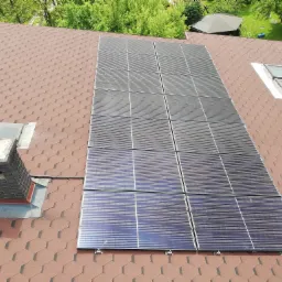 Panel fotowoltaiczny zamontowany na dachu pokrytym brązową dachówką bitumiczną, widok z góry, obok okna dachowe i komin.