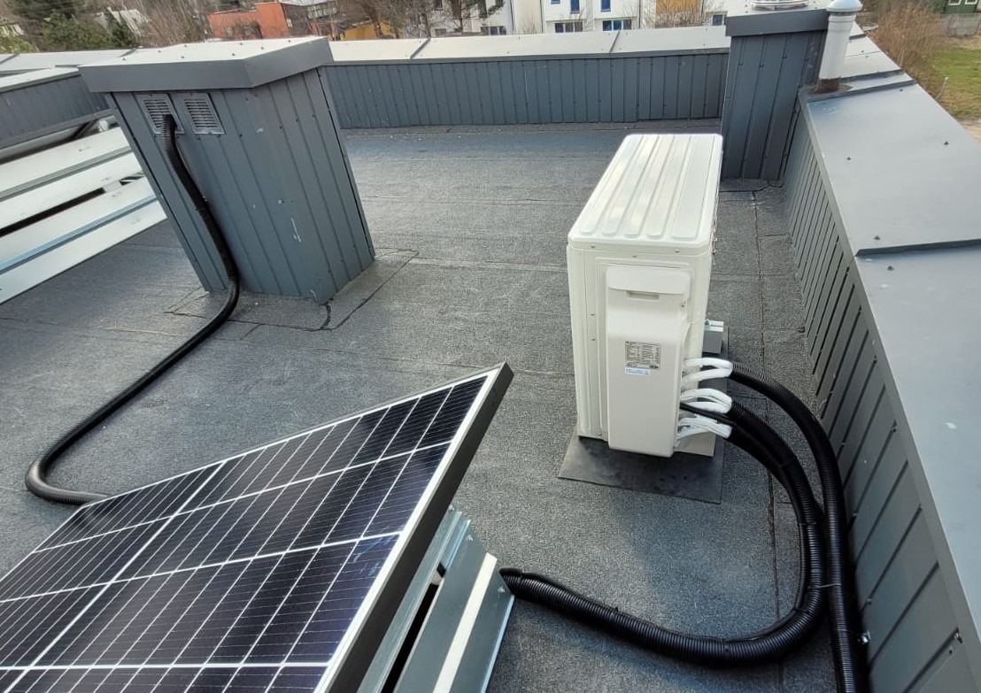 Instalacja klimatyzacji zewnętrznej na dachu płaskim, widoczne jednostka zewnętrzna, panele słoneczne i przewody instalacyjne.