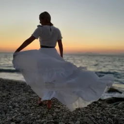 Kobieta w białej sukience wiruje na kamienistej plaży, plecami do aparatu, podczas zachodu słońca nad morzem.