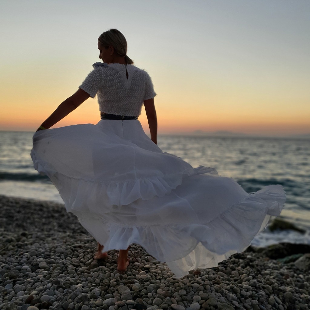 Kobieta w białej sukience wiruje na kamienistej plaży, plecami do aparatu, podczas zachodu słońca nad morzem.