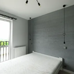 Minimalistyczna sypialnia z szarą tapetą o wyraźnej fakturze, białym łóżkiem bez pościeli, grzejnikiem pod oknem balkonowym z czarną balustradą i trzema lampami wiszącymi na długich kablach.