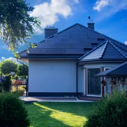 Dom jednorodzinny z panelami słonecznymi na dachu, widok z ogrodu w słoneczny dzień.