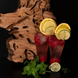 Dwa wysokie drinki z malinami i lodem, udekorowane plasterkami pomarańczy i limonki, na czarnym tle z drewnianą dekoracją i gałązką mięty