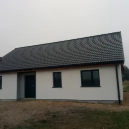 Parterowy dom szkieletowy z ciemną dachówką i czarną stolarką okienną, elewacja otynkowana na biało, widoczny fragment rynny spustowej.