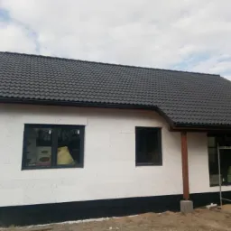 Niedokończony dom z białych bloczków z ciemną dachówką, widoczne rusztowania i okna z folią ochronną w środku.