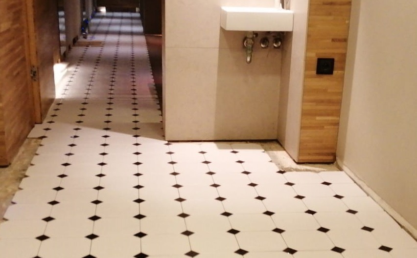 Ułożona podłoga z białych płytek z czarnymi rombami w korytarzu, widoczna umywalka i fragment drewnianej obudowy.