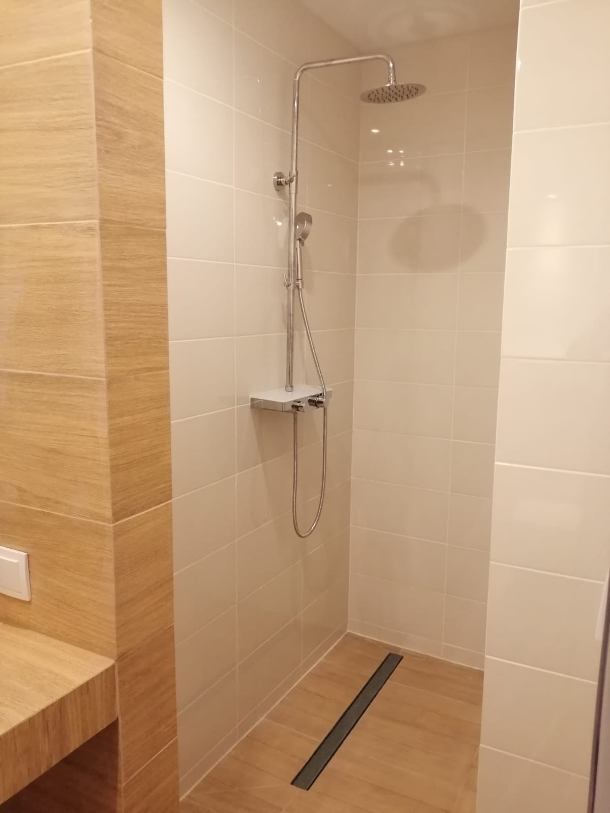 Nowoczesna kabina prysznicowa z deszczownicą i odpływem liniowym, wykończona beżowymi płytkami imitującymi drewno, z widocznym fragmentem blatu umywalki.