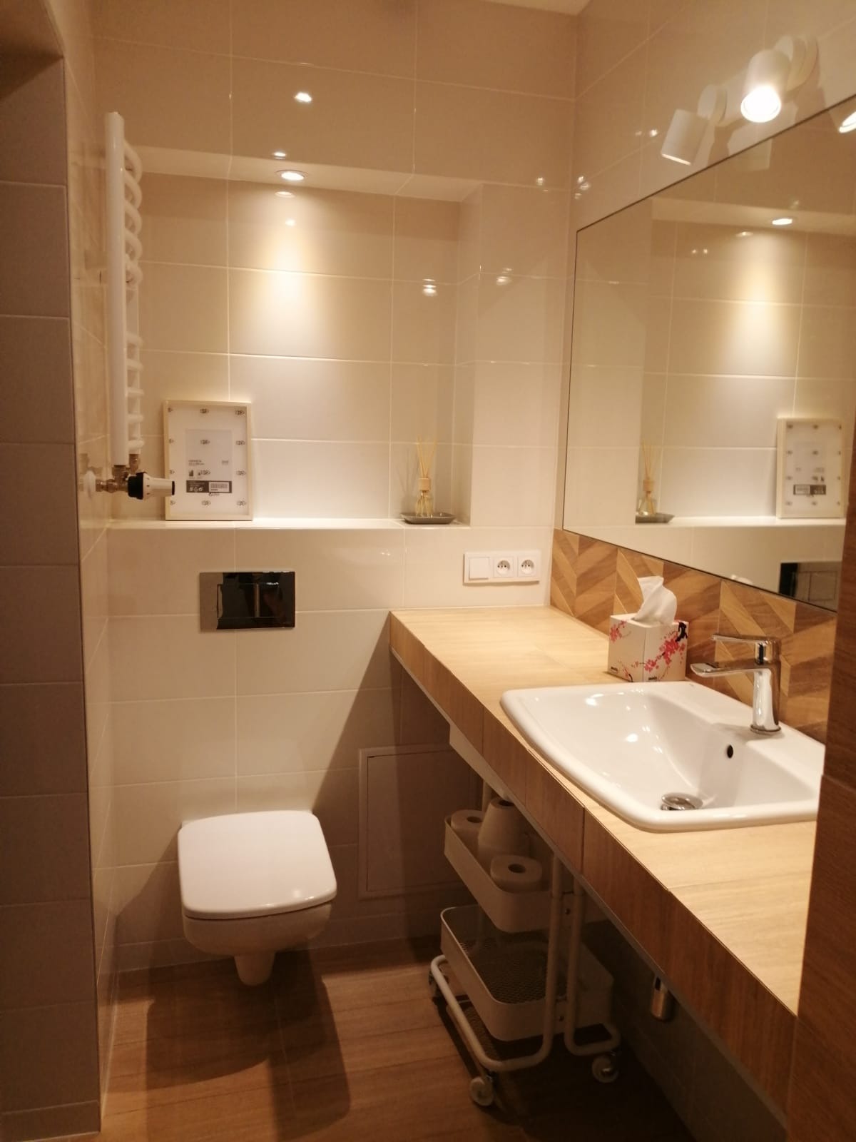 Jasna łazienka z beżowymi płytkami, drewnianym blatem pod umywalką, białym sedesem i grzejnikiem drabinkowym, oświetlona punktowymi lampami sufitowymi.