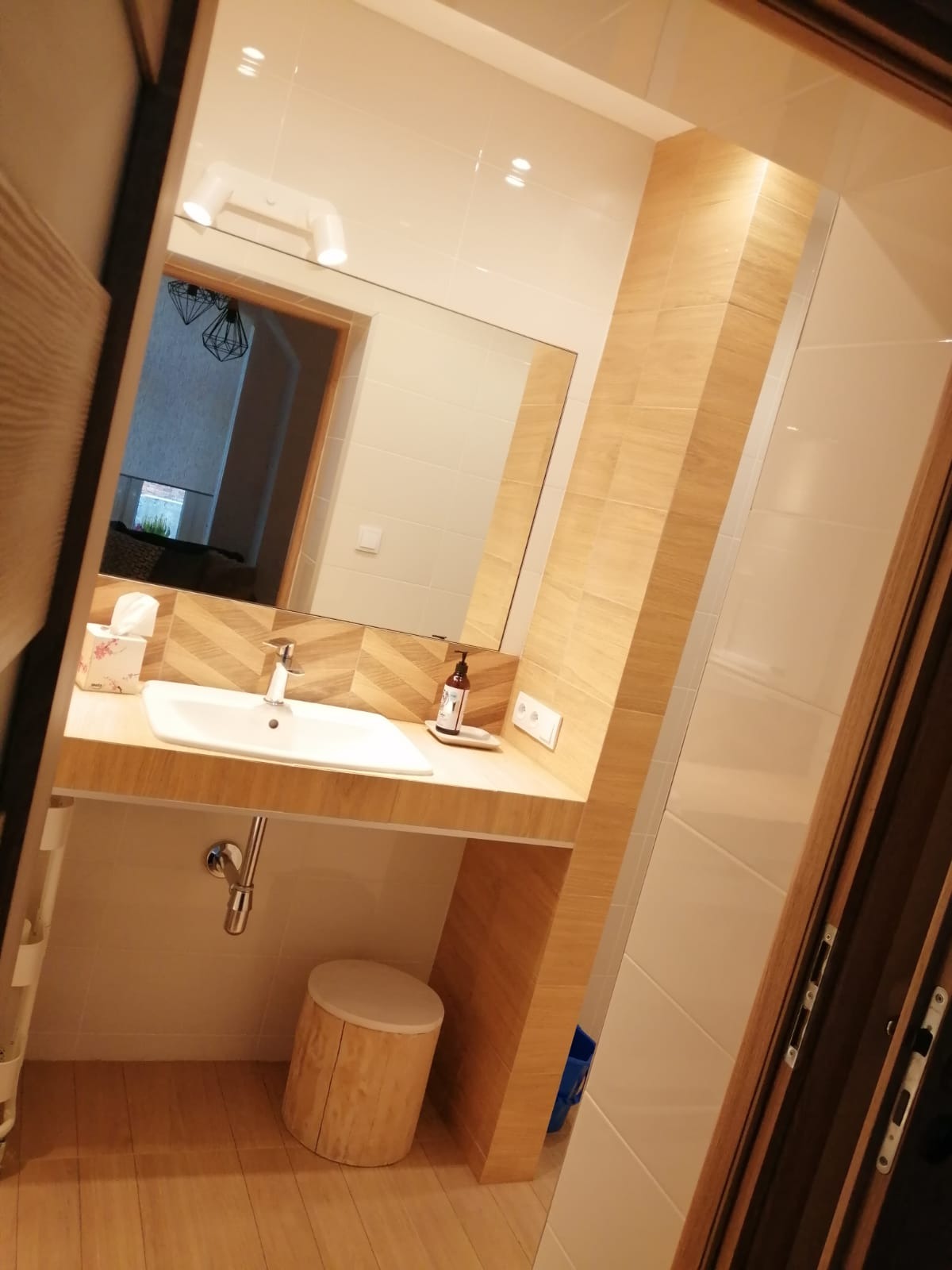 Nowoczesna łazienka po renowacji: umywalka nablatowa z baterią, lustro z podświetleniem, drewnopodobne płytki na ścianach i podłodze, oraz stołek z pnia drzewa.