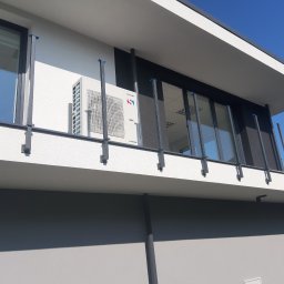 KrisCold - Biała jednostka zewnętrzna klimatyzatora Sinclair zamontowana na elewacji nowoczesnego budynku z elementami czerni i szarości, widoczna z dołu, z błękitnym niebem w tle.