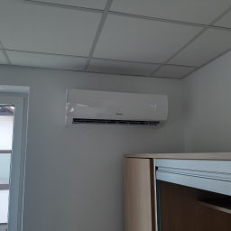 KrisCold - Biały klimatyzator Sinclair zamontowany na ścianie w jasnym pomieszczeniu z sufitem kasetonowym i częściowo widoczną szafą z przesuwnymi drzwiami.