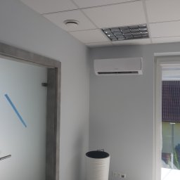 KrisCold - Klimatyzator ścienny zamontowany w biurze w Rzeszowie, widok na zewnątrz przez okno z roletami, obok drzwi z matową szybą i niebieską taśmą, w tle sufit kasetonowy z lampą.