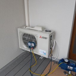 KrisCold - Zewnętrzna jednostka klimatyzacji Mitsubishi Electric zamontowana na ścianie budynku, podłączona do manometrów i pompy próżniowej podczas serwisu, widoczny tester szczelności.