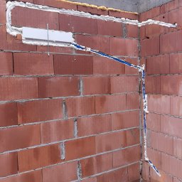 KrisCold - Montaż instalacji chłodniczej w nowym budynku: widoczne rury w izolacji piankowej prowadzone wzdłuż ceglanej ściany, z zamontowaną skrzynką przyłączeniową i metalową listwą montażową.