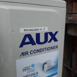 KrisCold - Boczny widok jednostki zewnętrznej klimatyzatora marki AUX z naklejką informacyjną 'Klimatyzator nr. 2' i logo firmy 'KRIS cold KLIMATYZACJA MONTAŻ - SERWIS' z numerem telefonu 502 560 020, na tle...