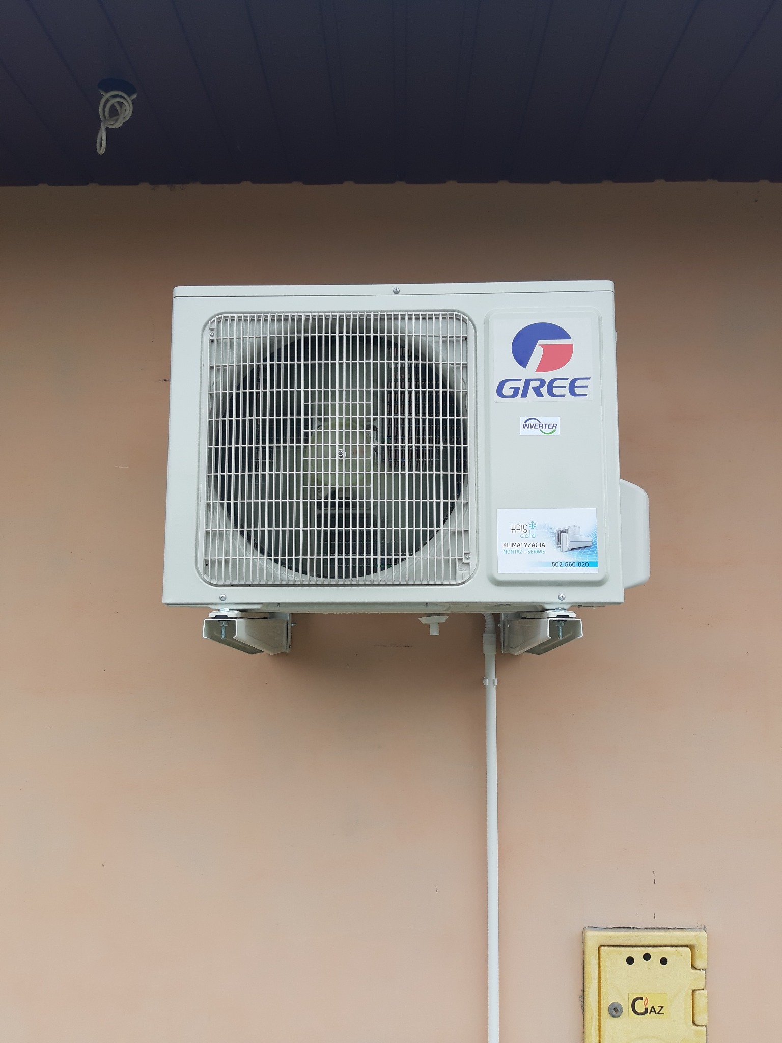 Zamontowana na zewnątrz jednostka klimatyzacji Gree z widoczną naklejką firmy instalacyjnej na tle beżowej ściany budynku.
