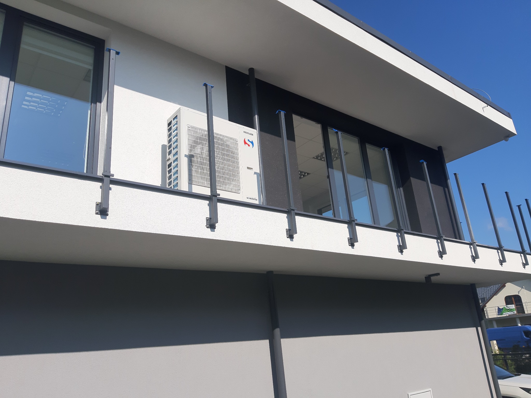 Biała jednostka zewnętrzna klimatyzatora Sinclair zamontowana na elewacji nowoczesnego budynku z elementami czerni i szarości, widoczna z dołu, z błękitnym niebem w tle.