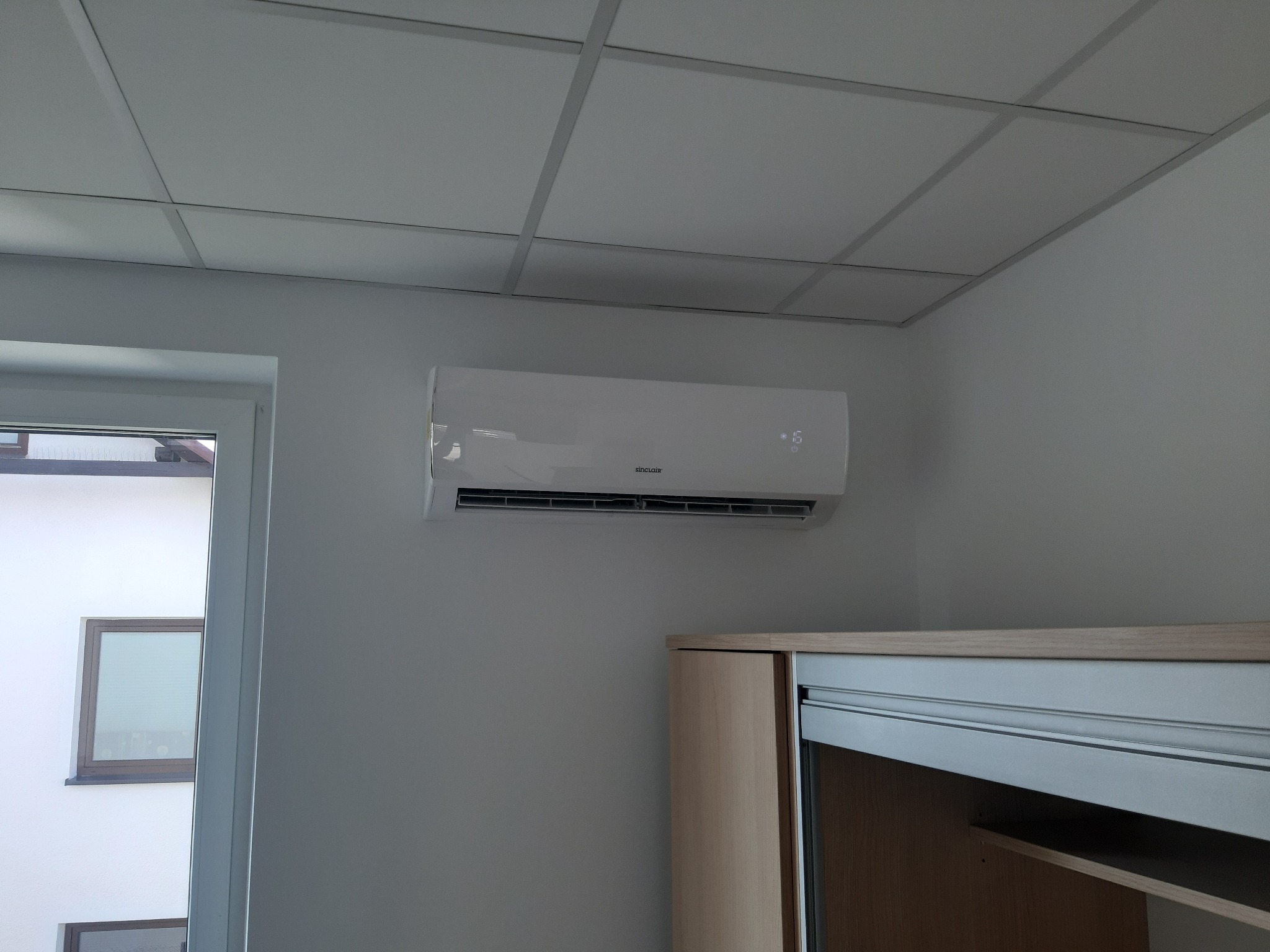 Biały klimatyzator Sinclair zamontowany na ścianie w jasnym pomieszczeniu z sufitem kasetonowym i częściowo widoczną szafą z przesuwnymi drzwiami.