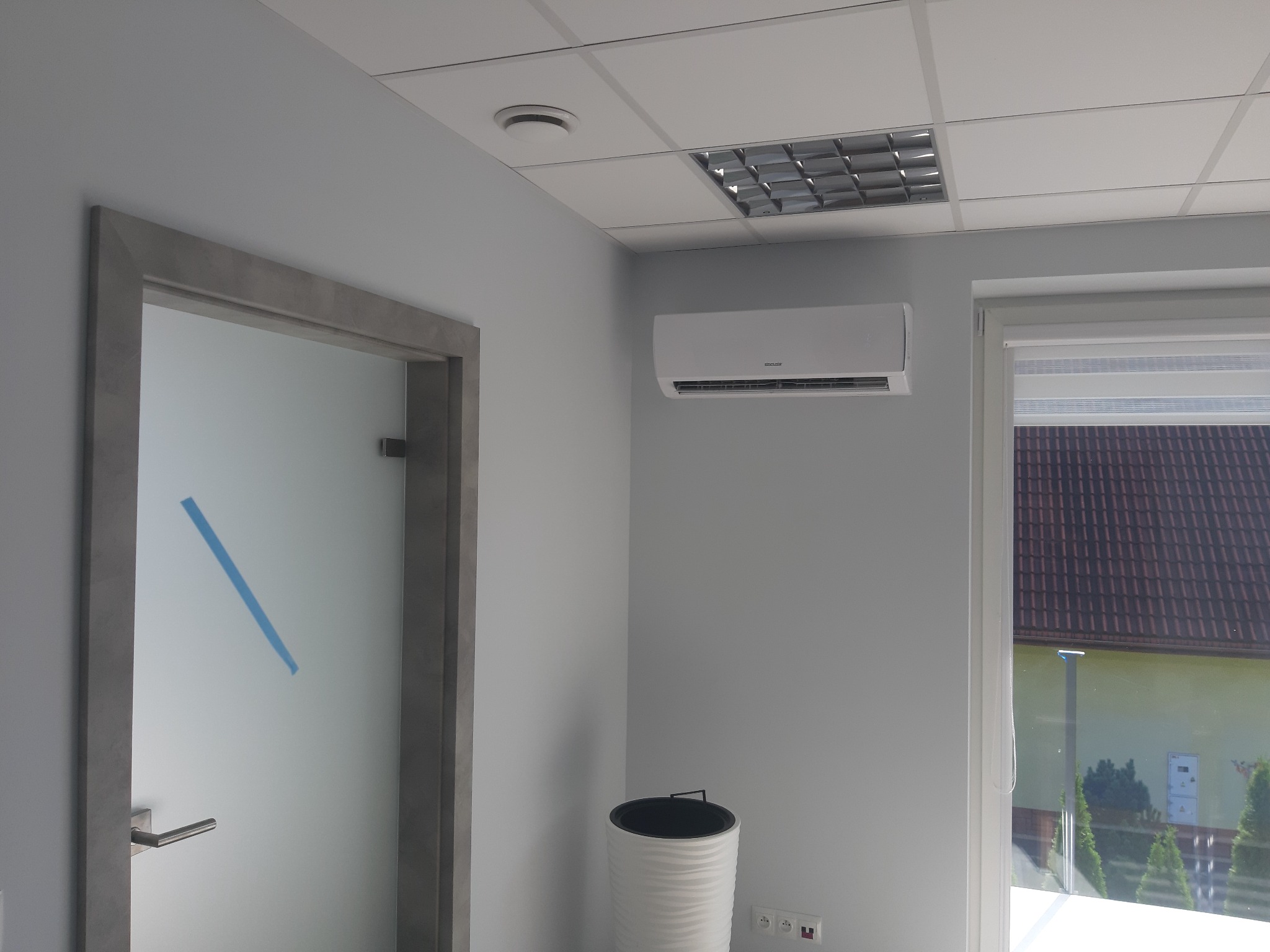 Klimatyzator ścienny zamontowany w biurze w Rzeszowie, widok na zewnątrz przez okno z roletami, obok drzwi z matową szybą i niebieską taśmą, w tle sufit kasetonowy z lampą.