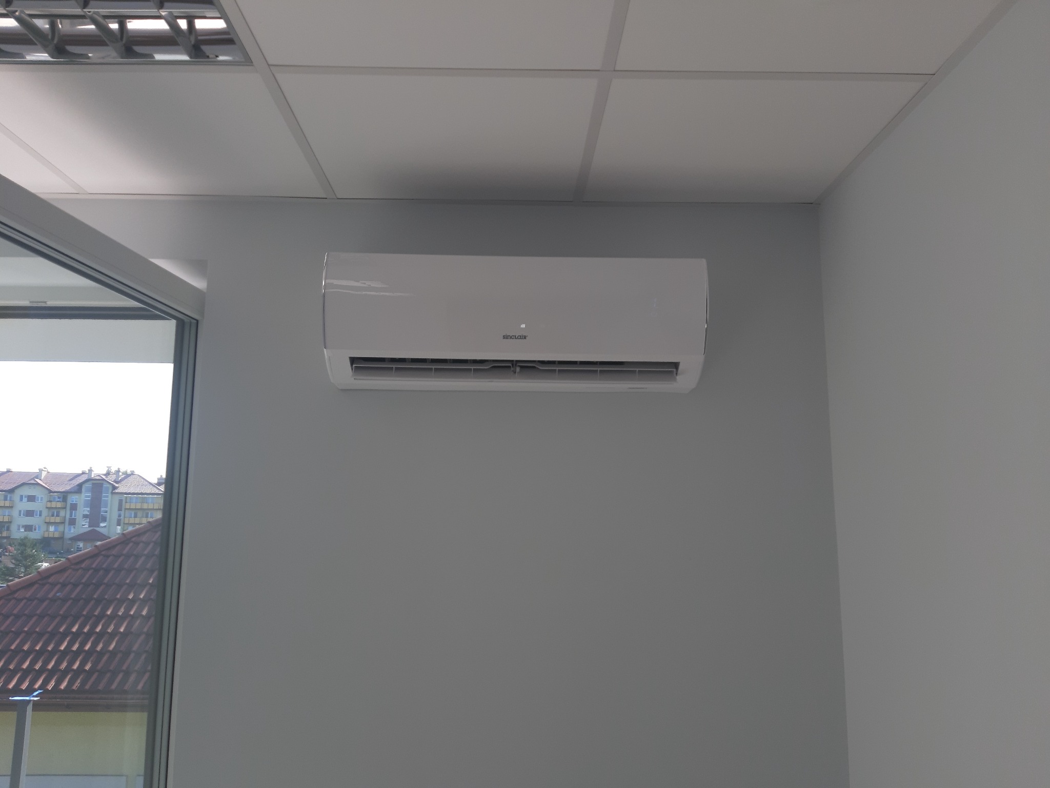 Biała jednostka klimatyzacyjna Sinclair zamontowana na szarej ścianie w biurze, widok przez okno na budynki mieszkalne.